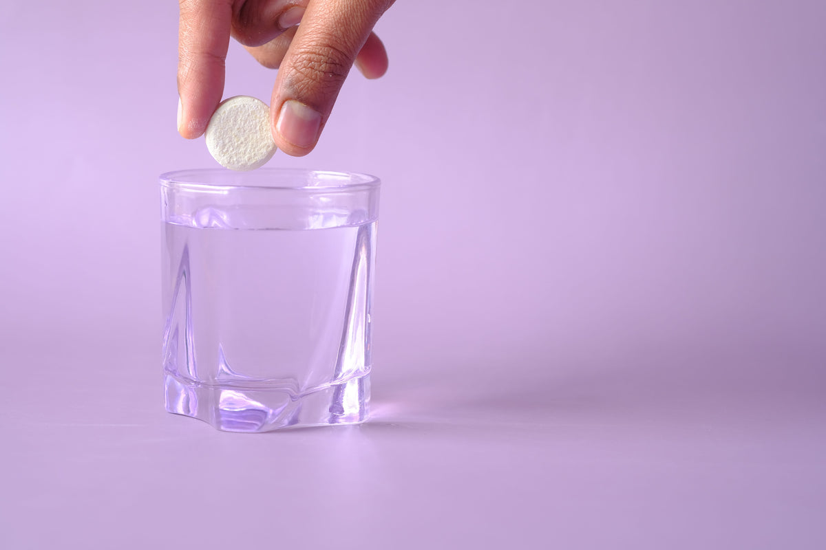 A hand dropping a pill into a water glass with a purple backdrop