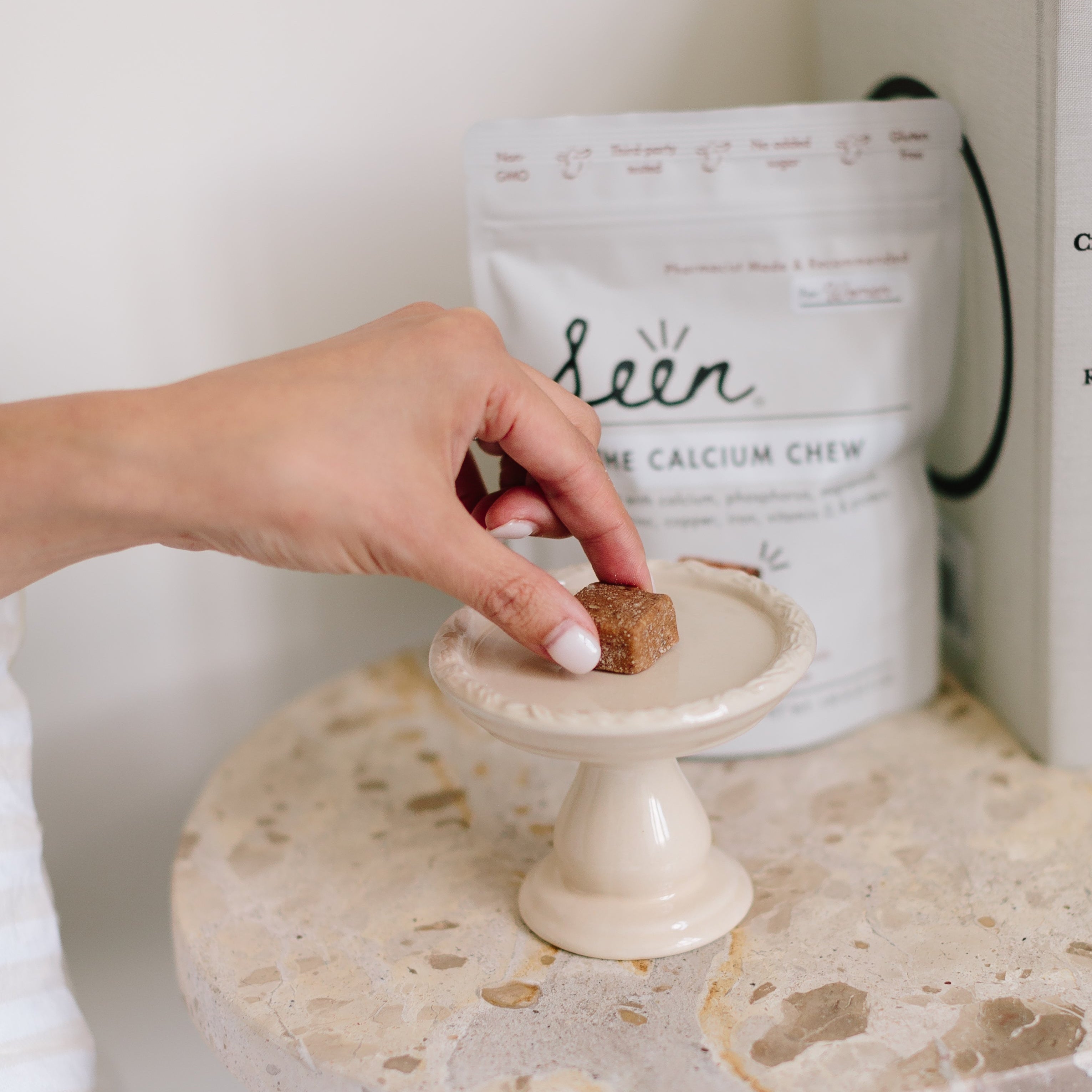 Person grabbing a Seen Calcium Supplement off a bedside table with a bag of 'Seen Calcium Chews' in the background.
